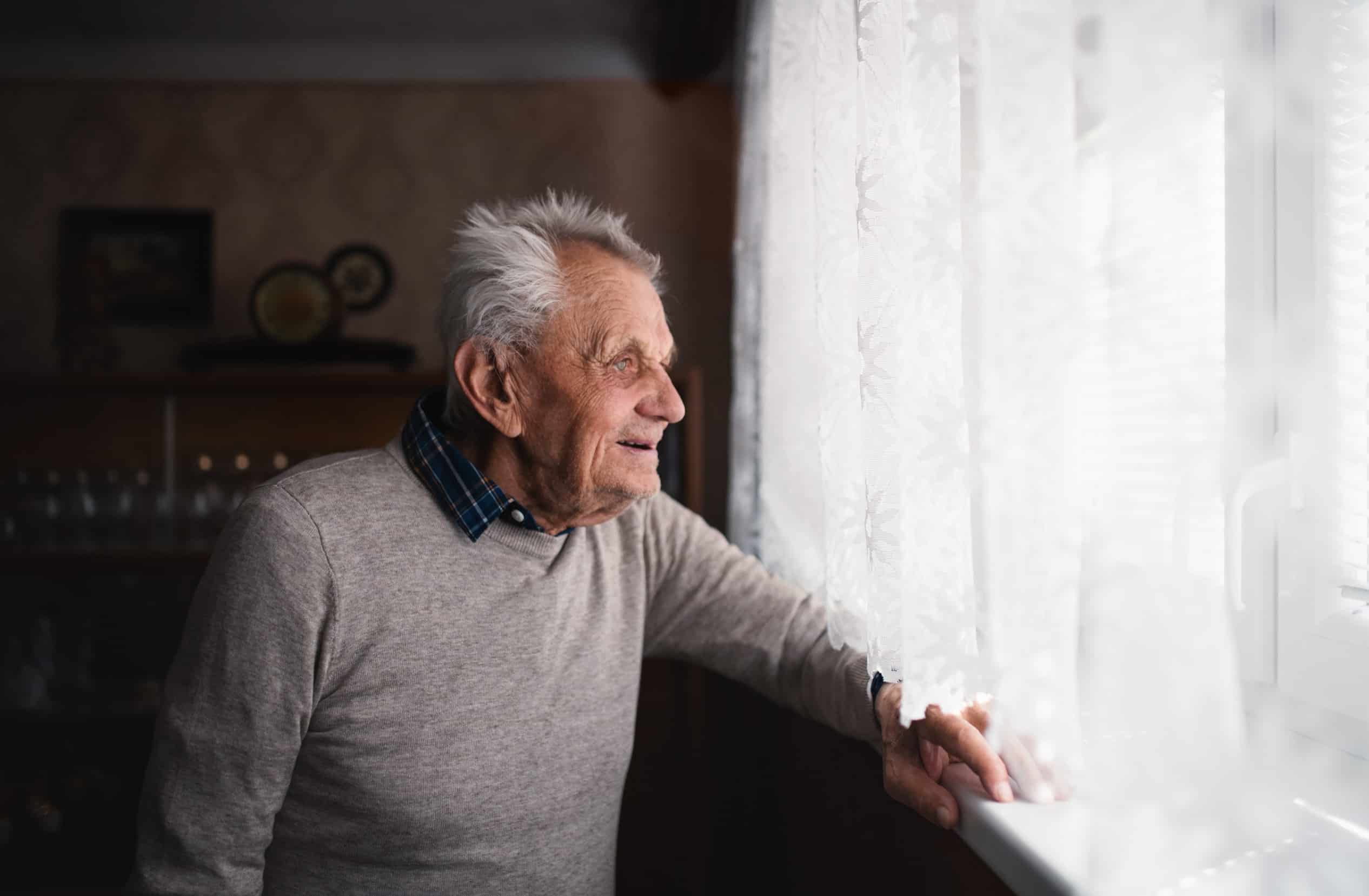 Photograph of elderly man standing indoors at home, looking out through window
