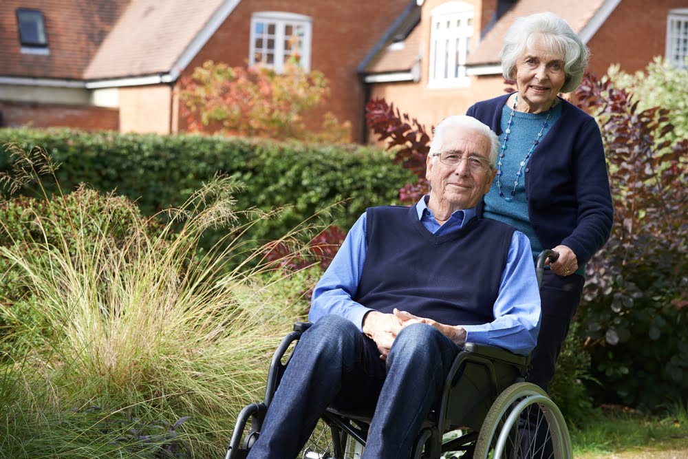 Elderly woman standing behind elderly man who is seated in a wheelchair
