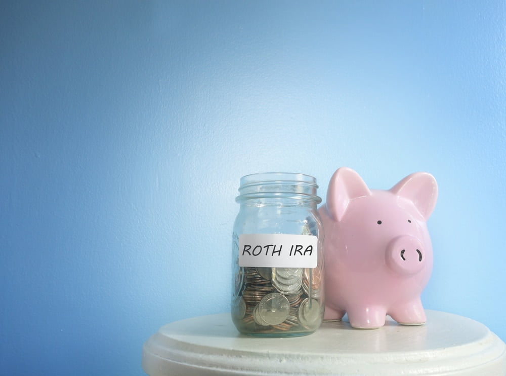 Jar containing money labeled Roth IRA sitting on a table next to a piggy bank in front of a blue background